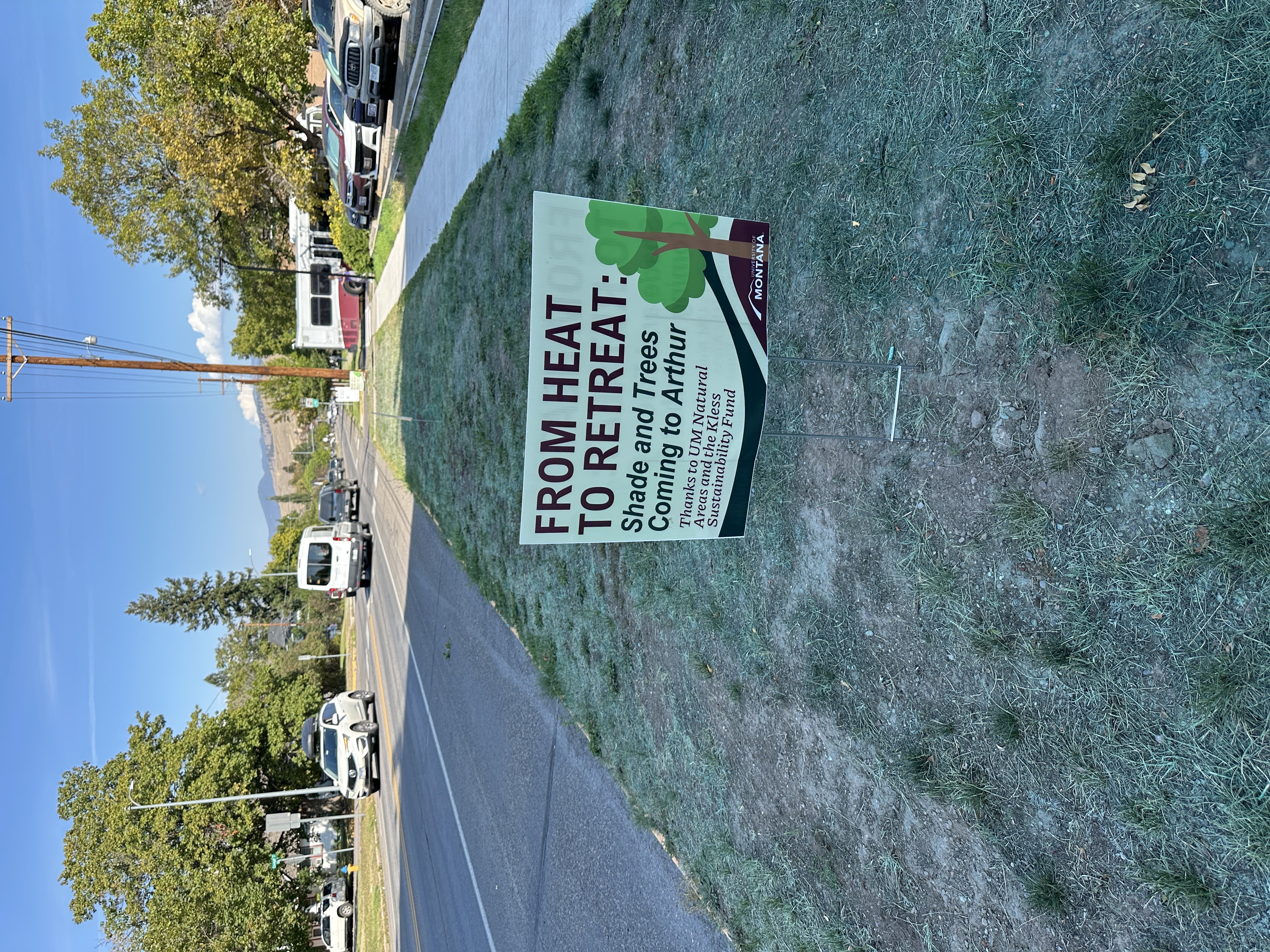 Yard sign on a grassy roadside reading “From Heat to Retreat: Shade and Trees Coming to Arthur,” with traffic and trees in the background.