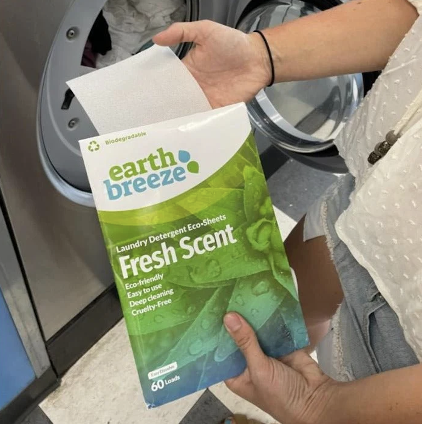 Hand holding an Earth Breeze Fresh Scent laundry detergent eco-sheets package and a sheet in front of an open washing machine.