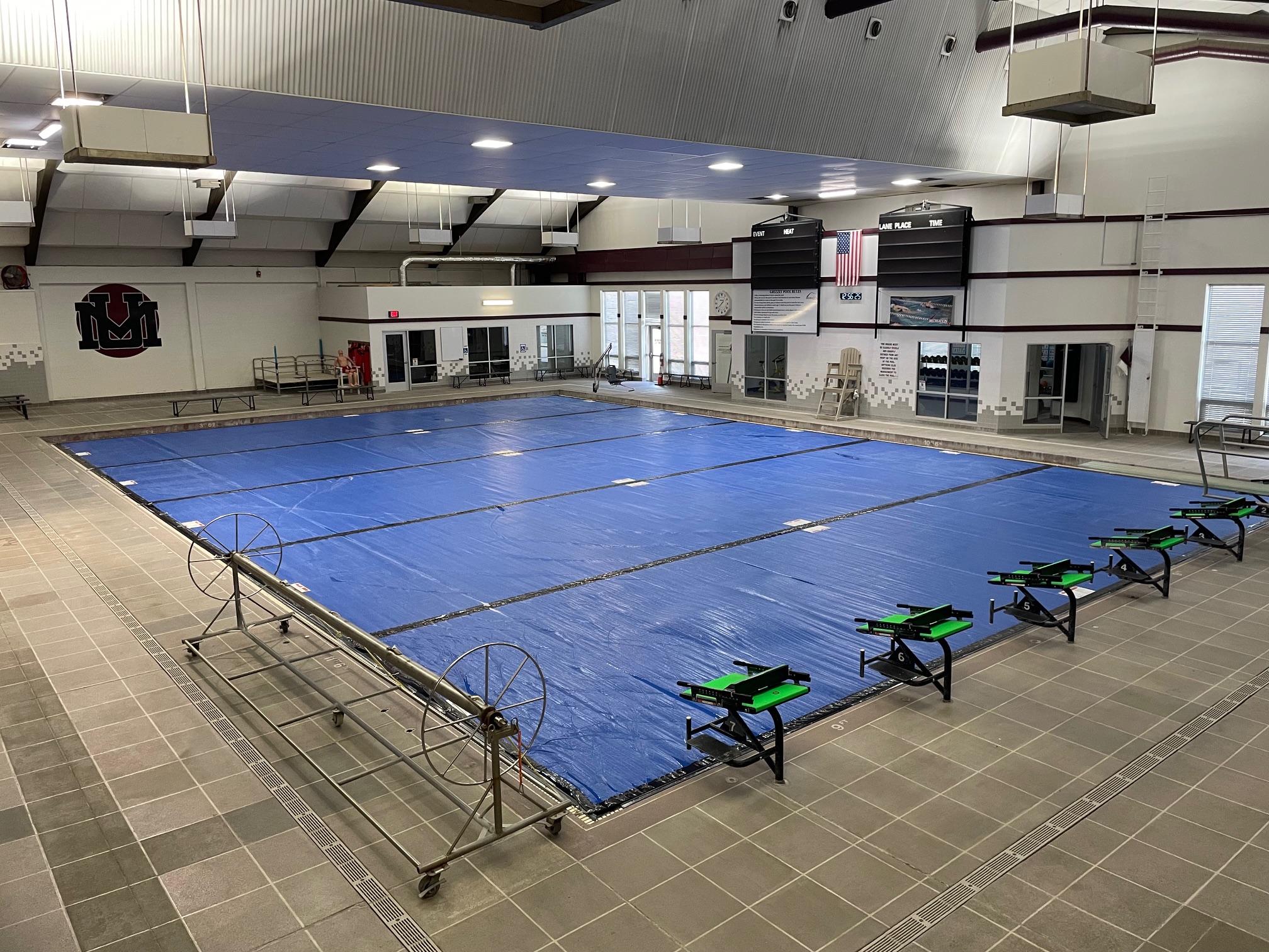 Indoor swimming pool covered with a blue safety cover, with starting blocks and a scoreboard visible.