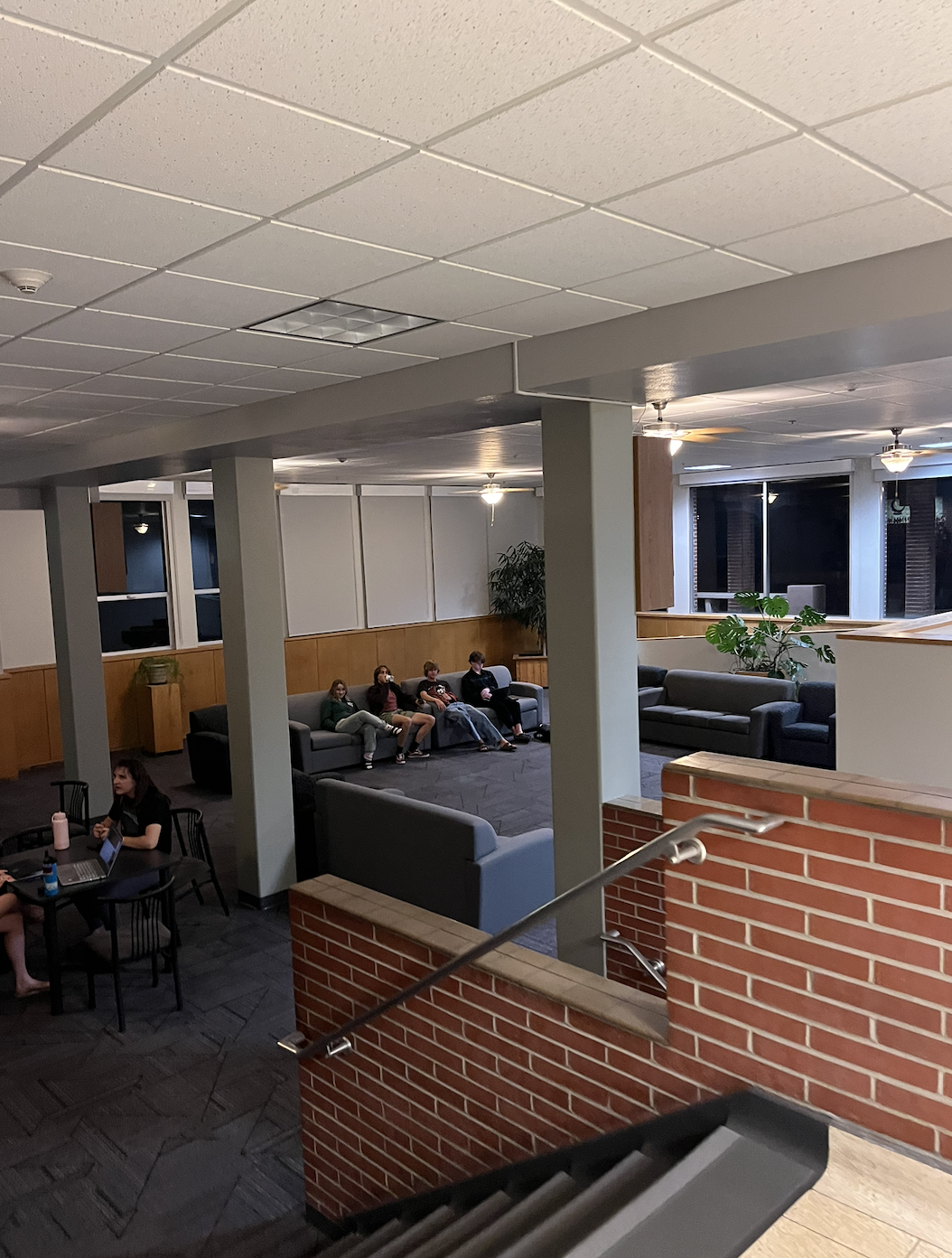 Student lounge area with couches and tables, with people sitting and studying near a stairwell.
