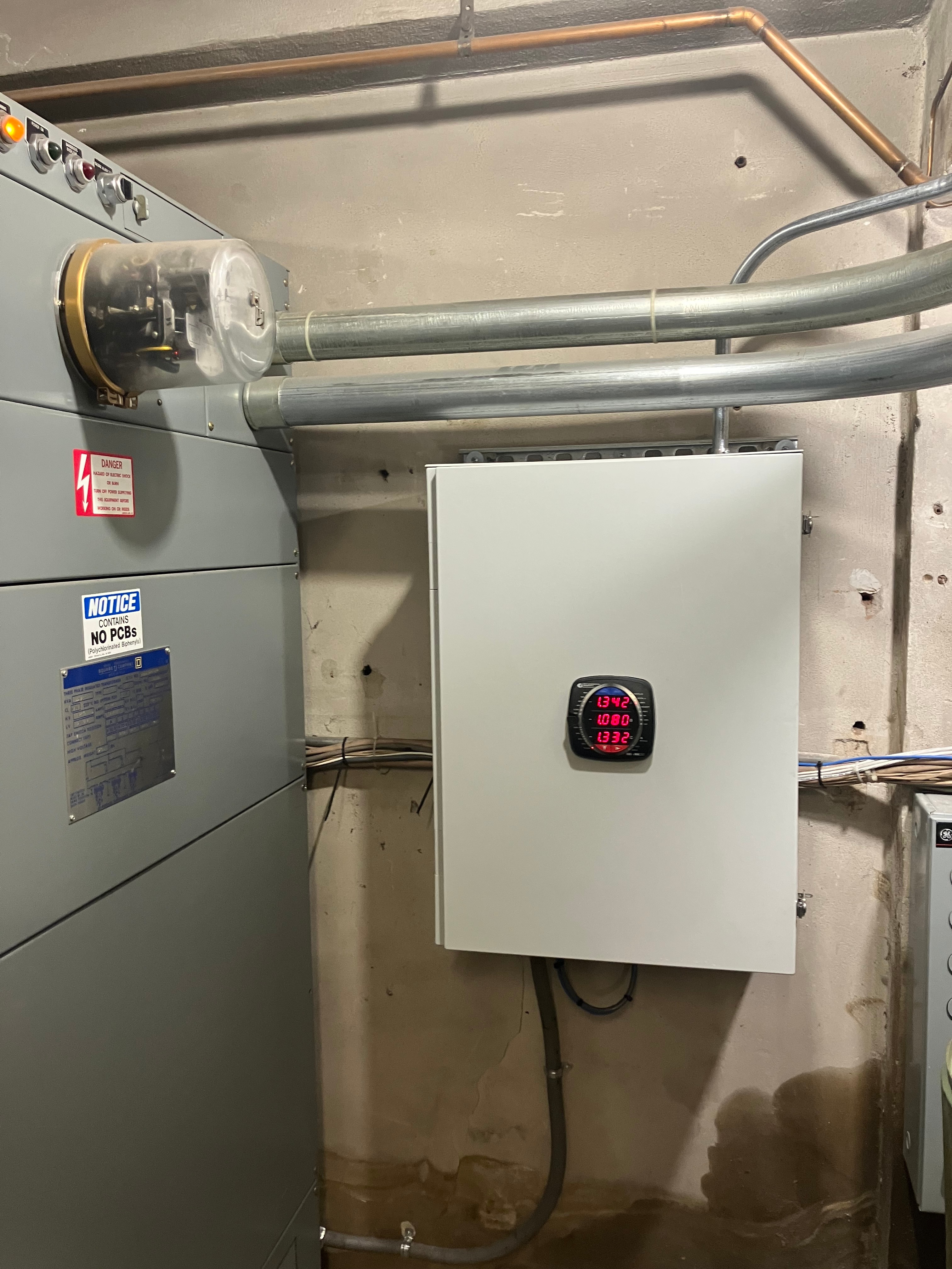 Wall-mounted electrical control panel with a digital display, conduit piping, and nearby utility equipment.