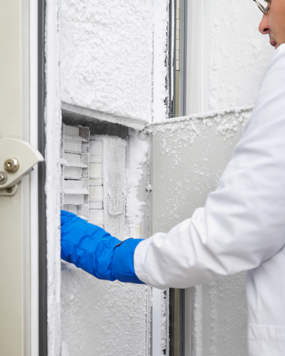 A scientist in a lab coat and blue gloves is retrieving items from a frost-covered freezer.