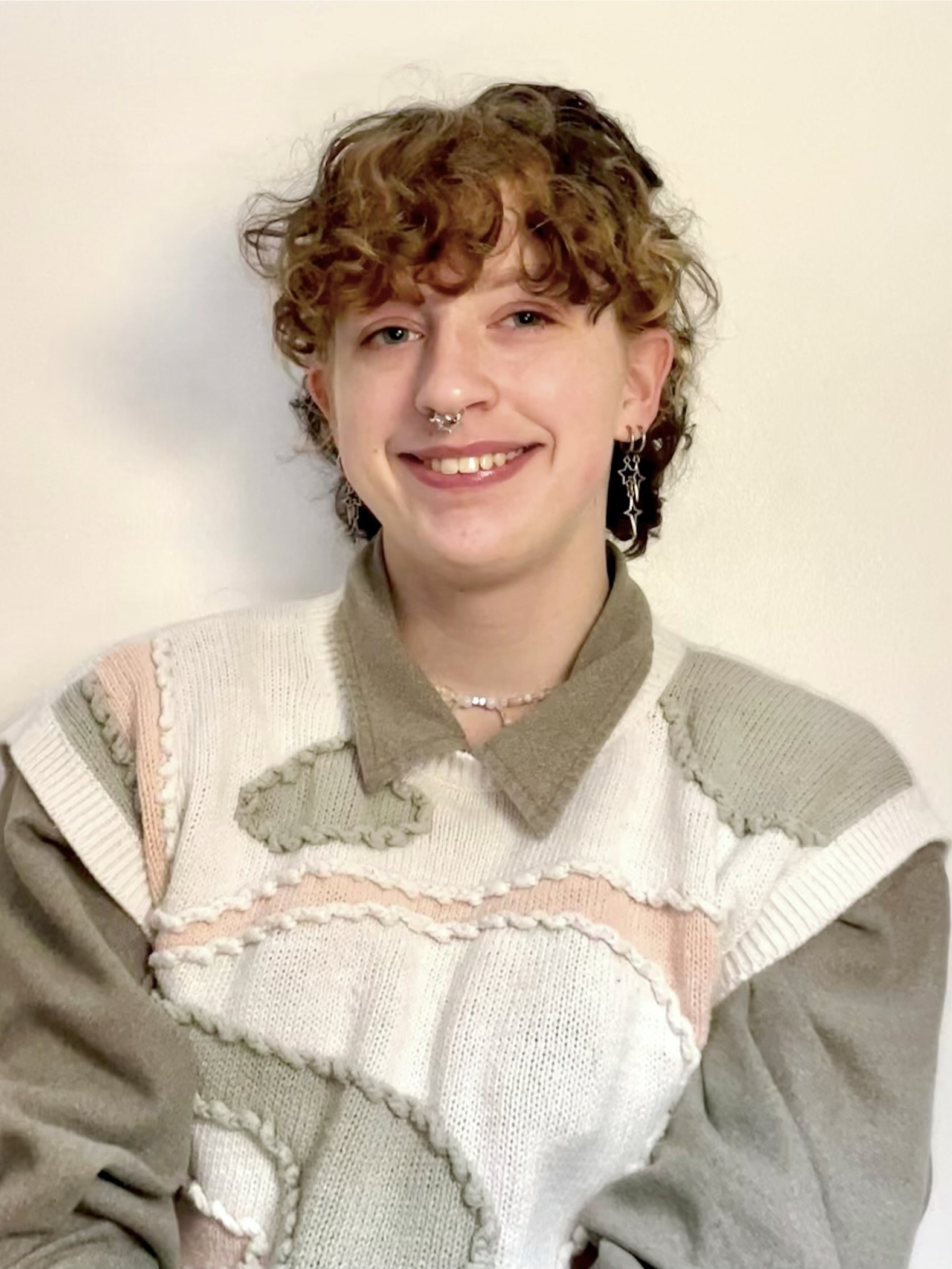 George smiling, curly blonde/light brown hair, nose and ear piercing, weating a white, pink, and brown sweater