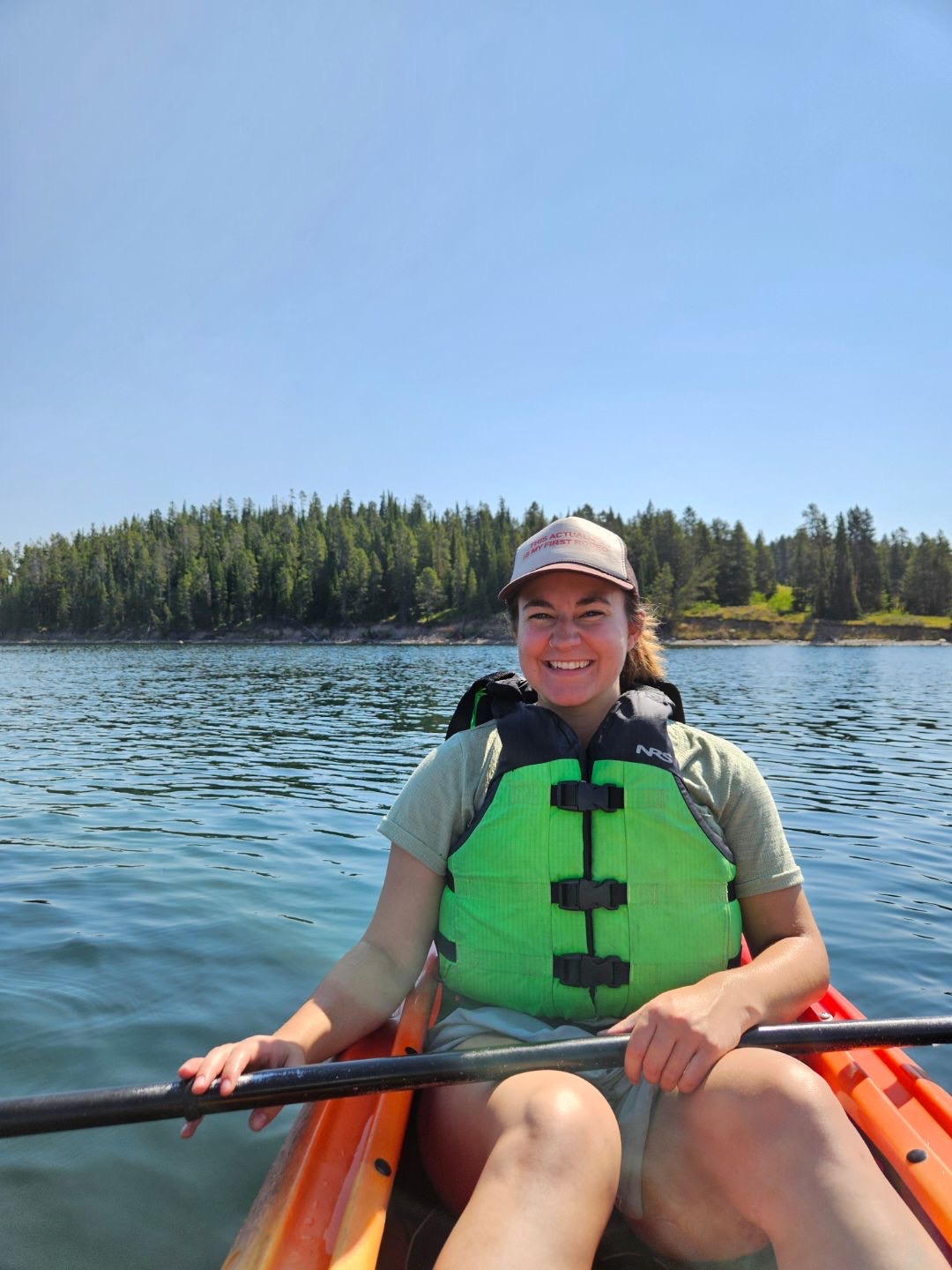 Julia is on an orange kayak and wearing and lime green life vest. She is smiling with evergreen trees behind her.