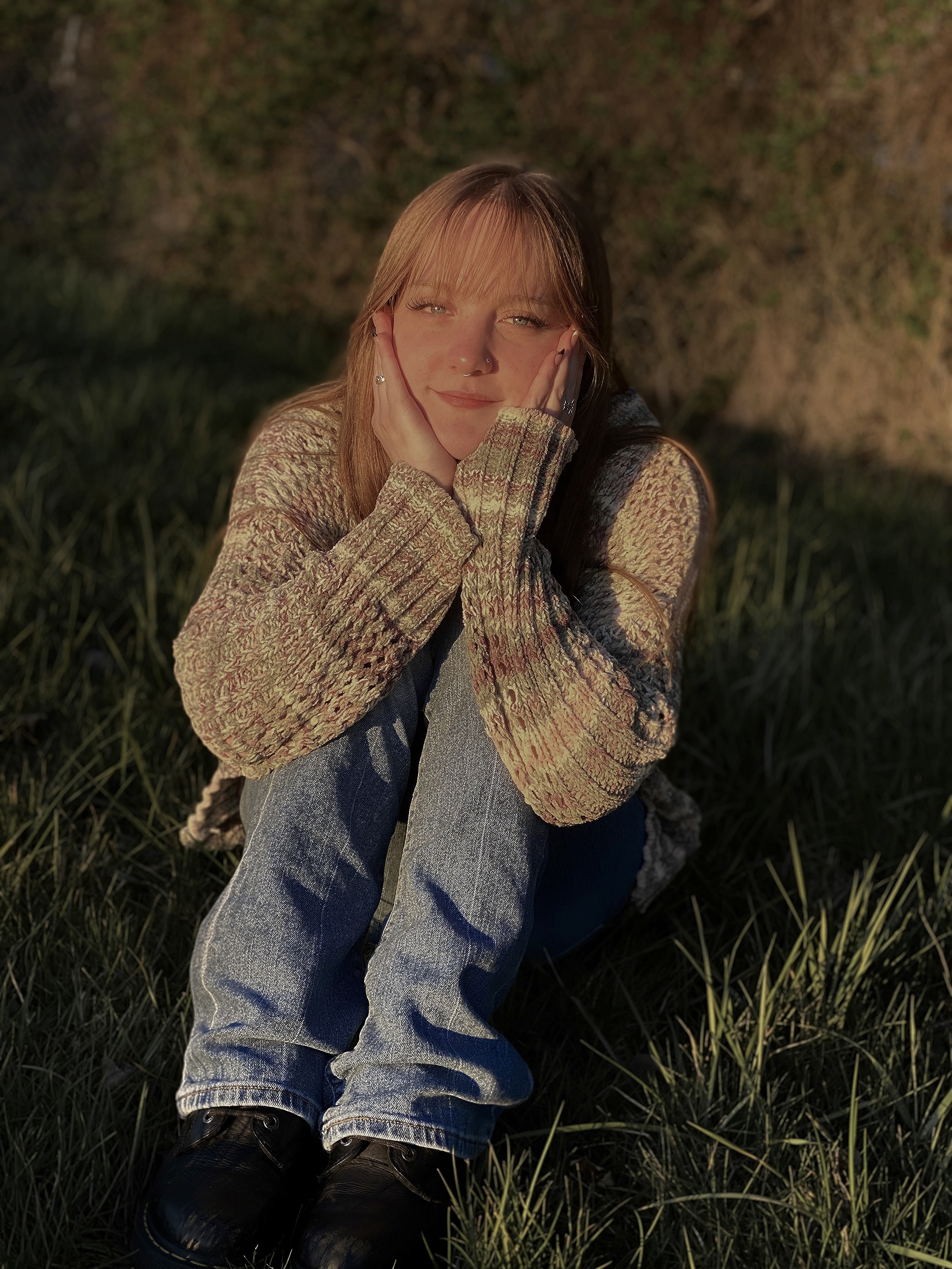 Image of Arianna smiling with hands on her face, she has straight blonde hair with bangs and is sitting on the ground wearing jeans and a sweater