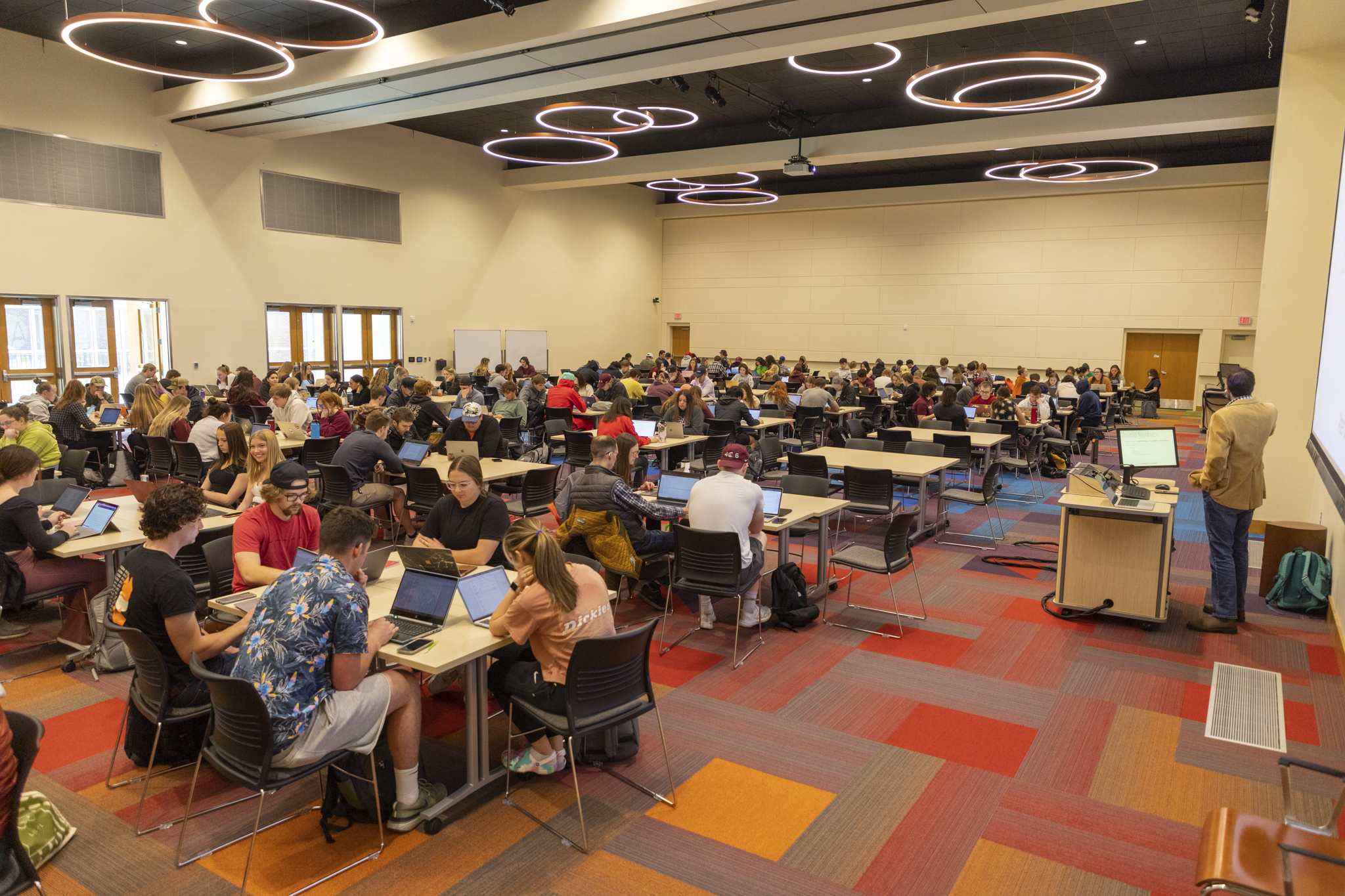 Large classroom of students sitting in a groups listening to the professors lecture. 