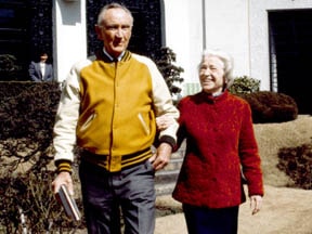 A vintage photograph of a couple walking together, with the man wearing a yellow and brown varsity jacket and the woman in a red coat, smiling at each other.
