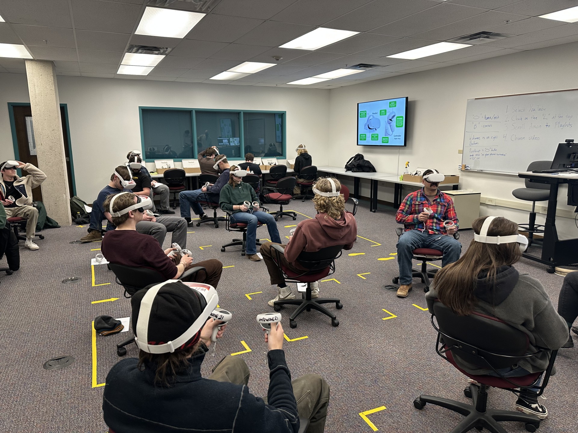 Image showing 12 students wearing VR headsets seated in chairs inside of the Buckhous Immersive Learning Space at the Mansfield Library. Visible in the background is an LCD display showing VR controls and a whiteboard with instructions written on it. . 