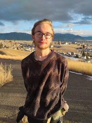 A young man wearing glasses and a patterned sweater stands on a hillside with a scenic view in the background.