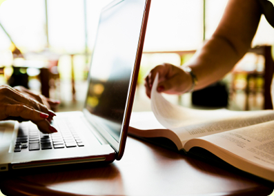 closeup view of hands typing on a laptop and another pair of hands leafing through a book