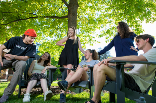 Students gathered outside on a sunny day under the shade of vibrant green leafy trees. 