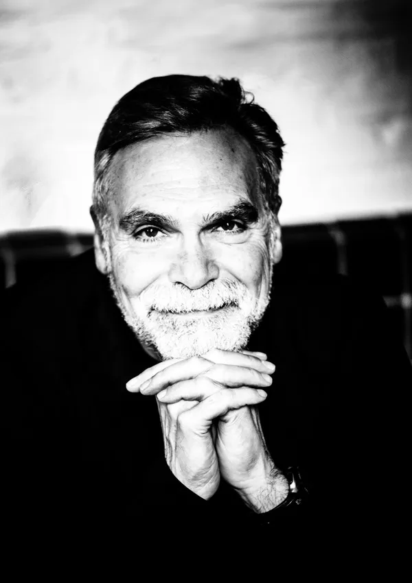 A person with salt and pepper hair rests his chin on clasped hands and smiles warmly at the camera.