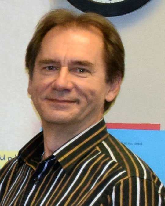 Smiling chris bujak in a striped button-down shirt, posing indoors against a wall with colorful papers in the background.