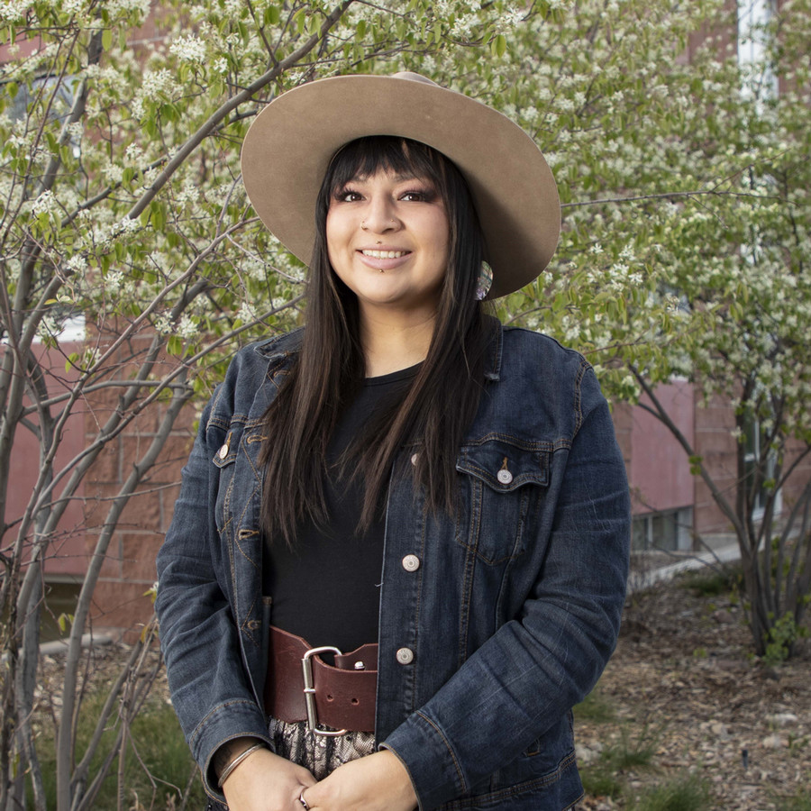 Courtney Little Axe wearing a denim jacket and wide-brimmed hat, smiling in front of flowering trees.