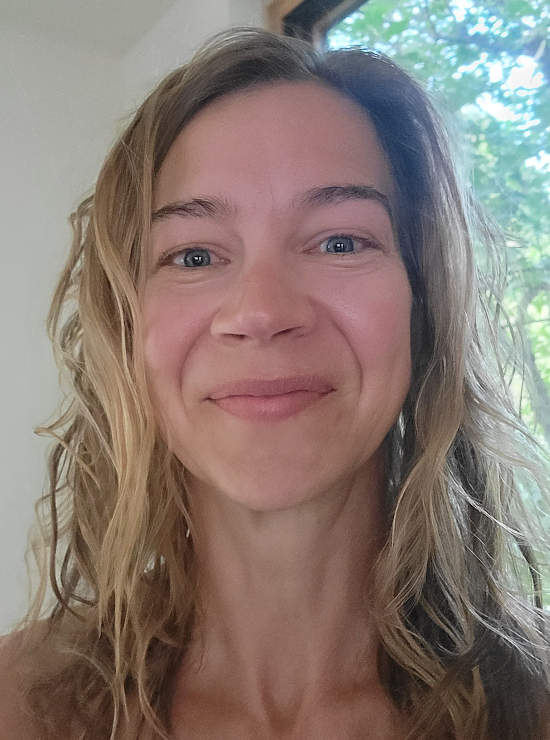 A smiling woman with wavy hair, standing in a bright space surrounded by greenery.