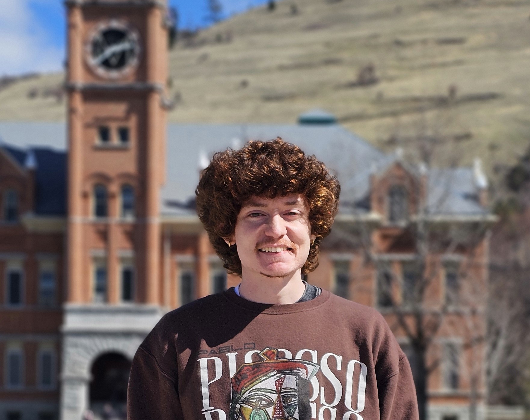 A young man with curly hair wearing a Picasso-themed sweatshirt stands in front of a historic building on a sunny day.