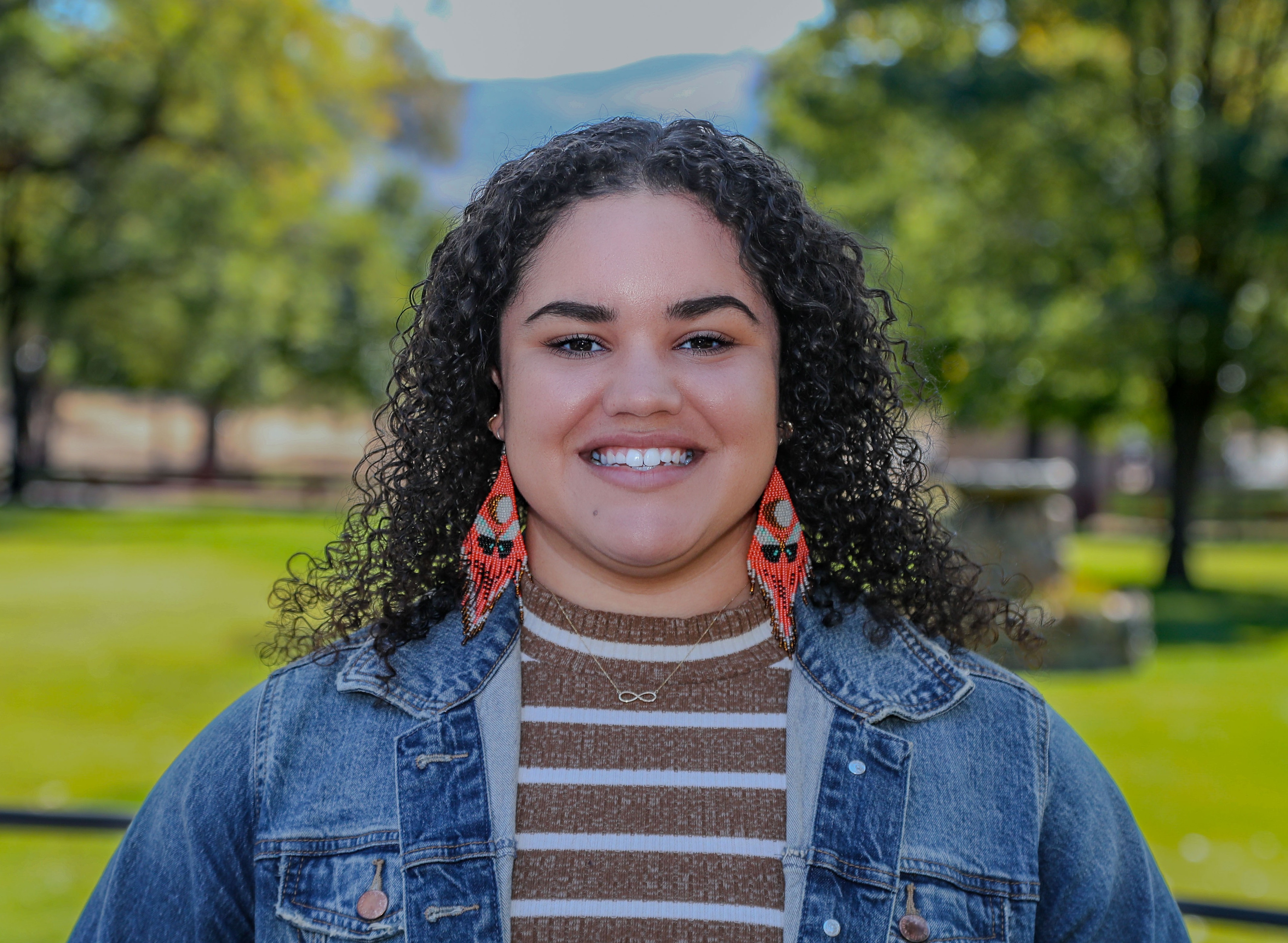 A smiling woman with curly hair wearing a denim jacket and colorful earrings stands outdoors with green trees in the background.