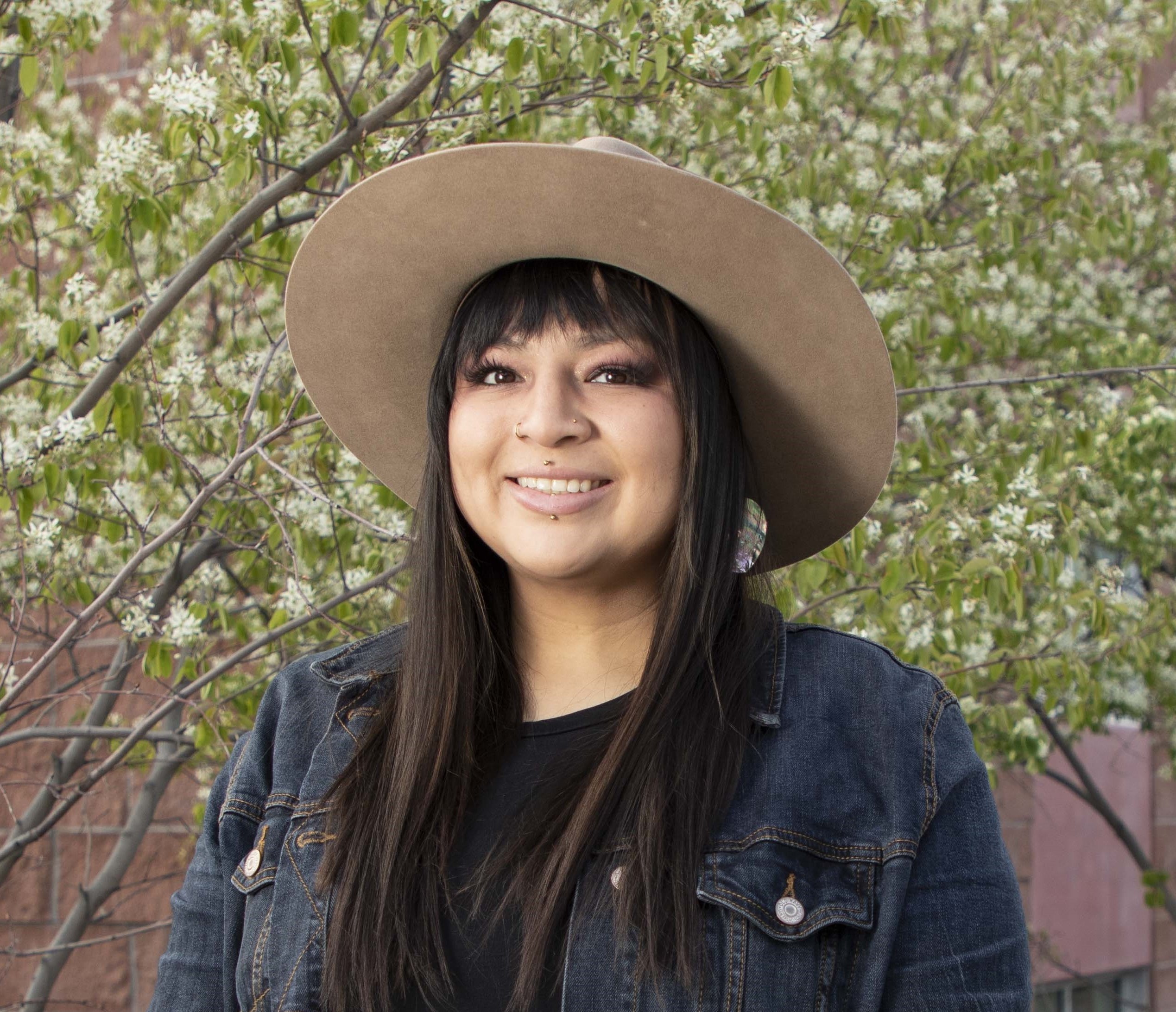 A woman wearing a denim jacket and wide-brimmed hat stands in front of blooming trees, smiling confidently.