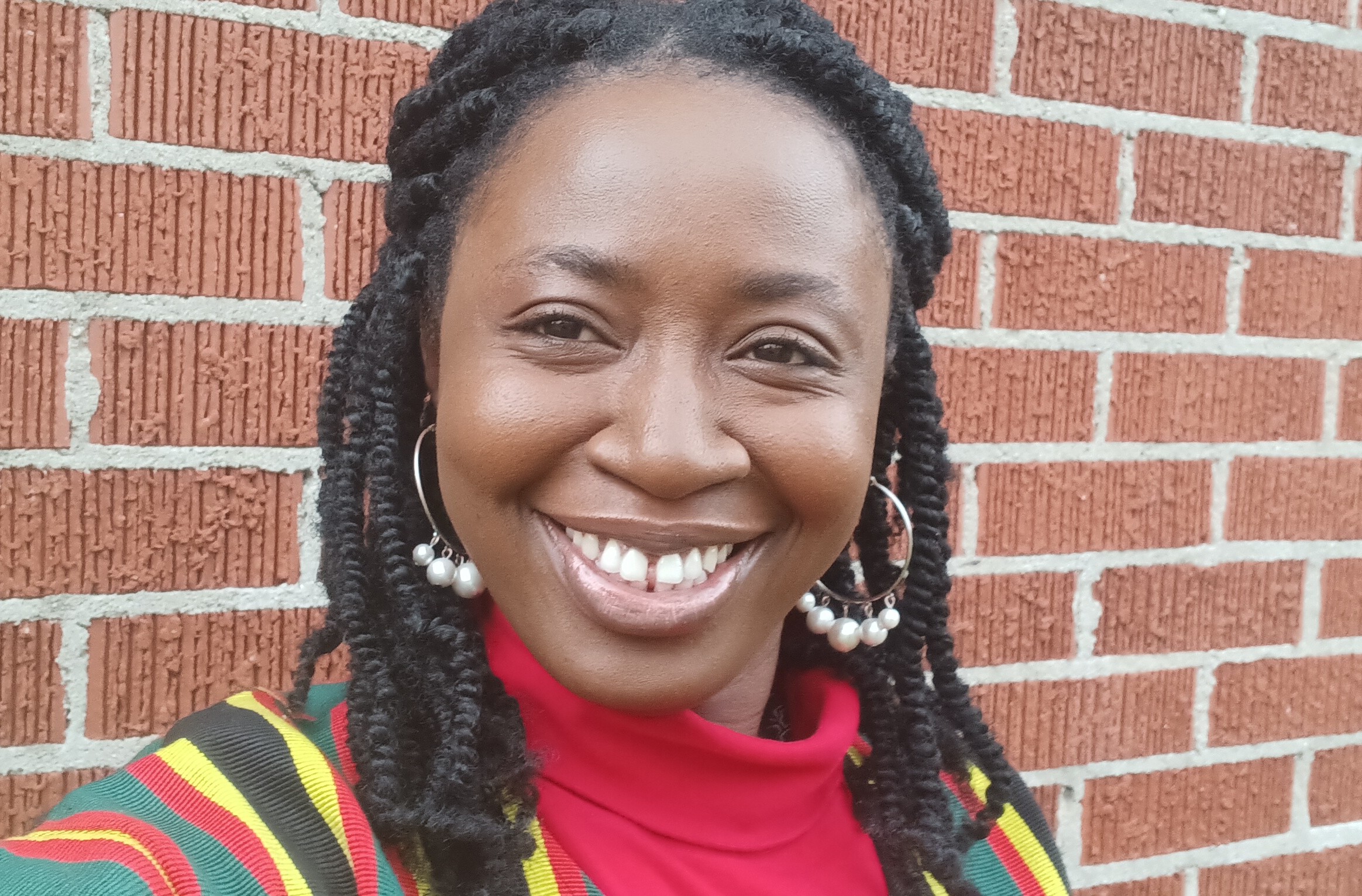 A smiling woman with natural hair poses in front of a brick wall, wearing a colorful patterned shawl and pearl earrings.