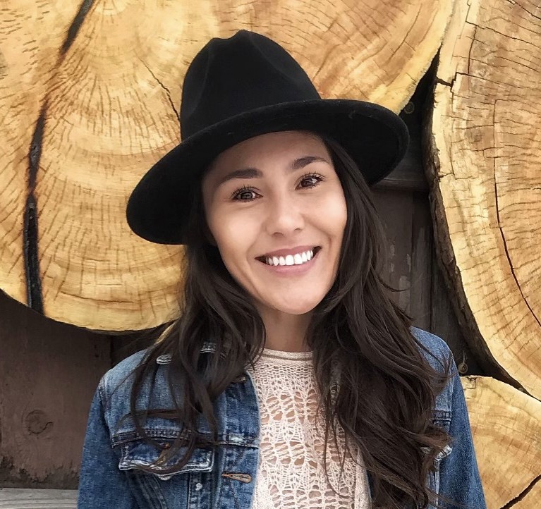 A smiling woman wearing a black hat and denim jacket stands in front of a wooden backdrop.