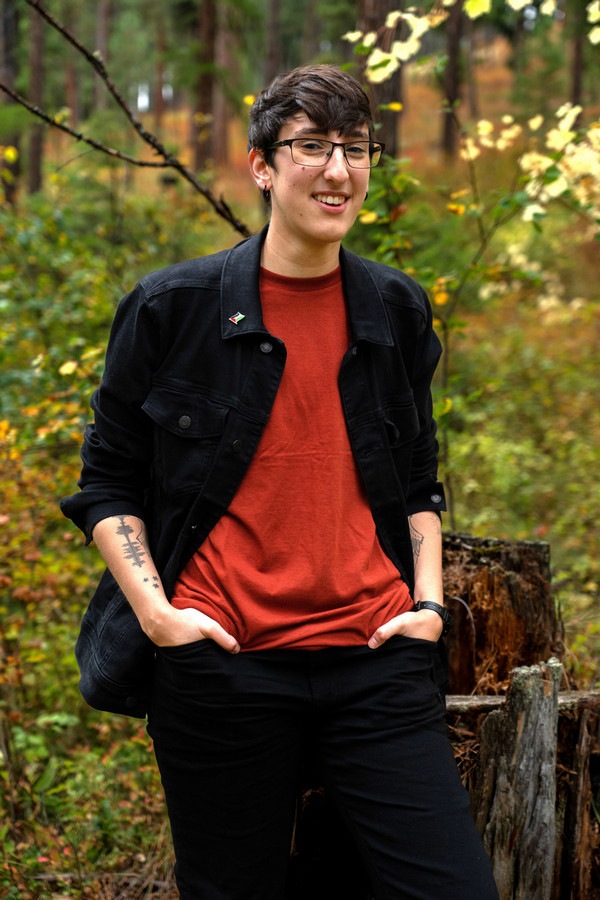 Nas Jabr stands in a forest, wearing a black jacket over a red shirt, with a relaxed smile.