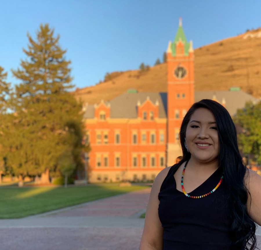A smiling Shalbilyn Tall Whiteman stands in front of historic University Hall, surrounded by greenery and hills