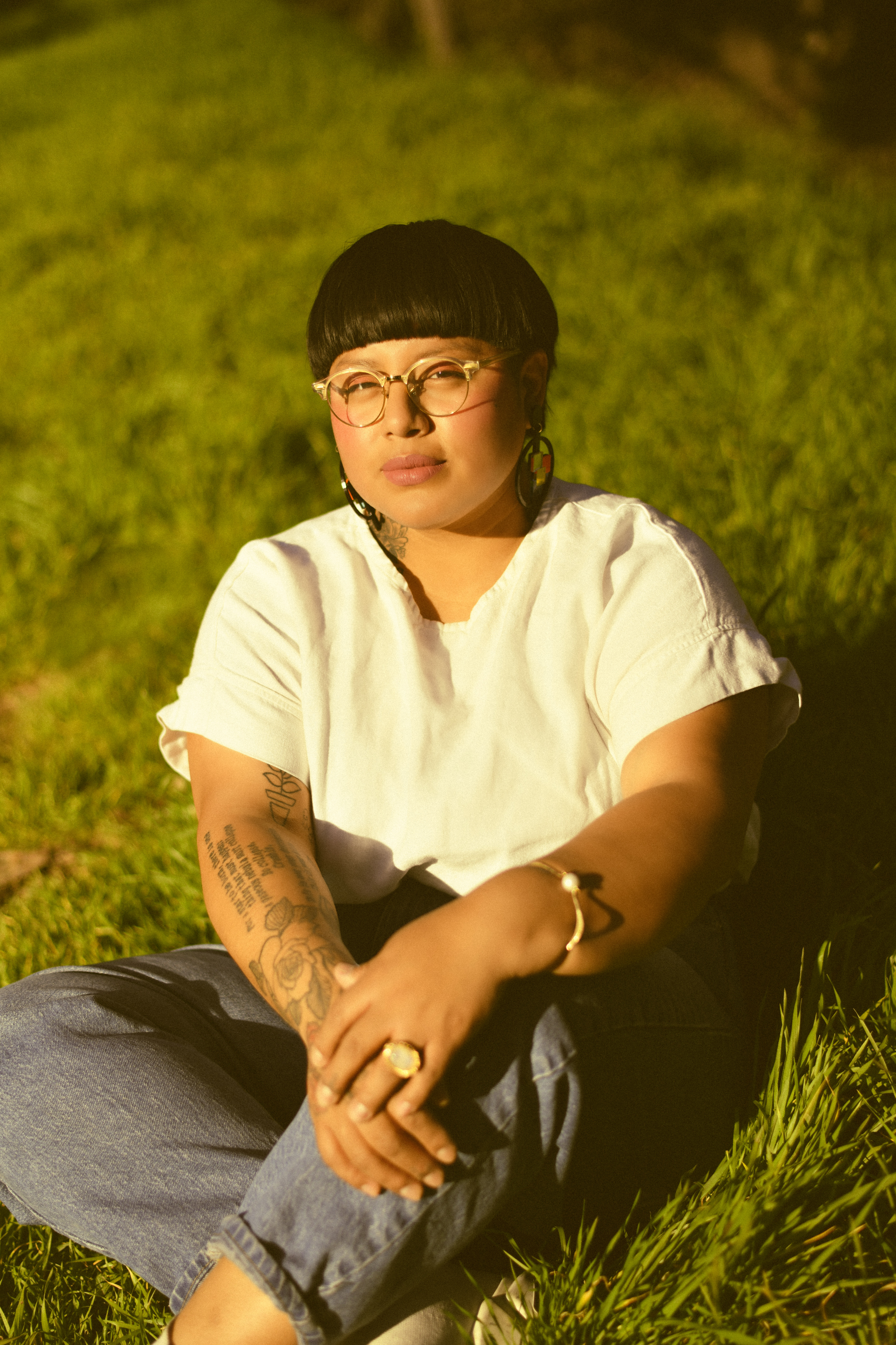 A woman with a stylish haircut and glasses sits on grass, wearing a casual outfit with visible tattoos.