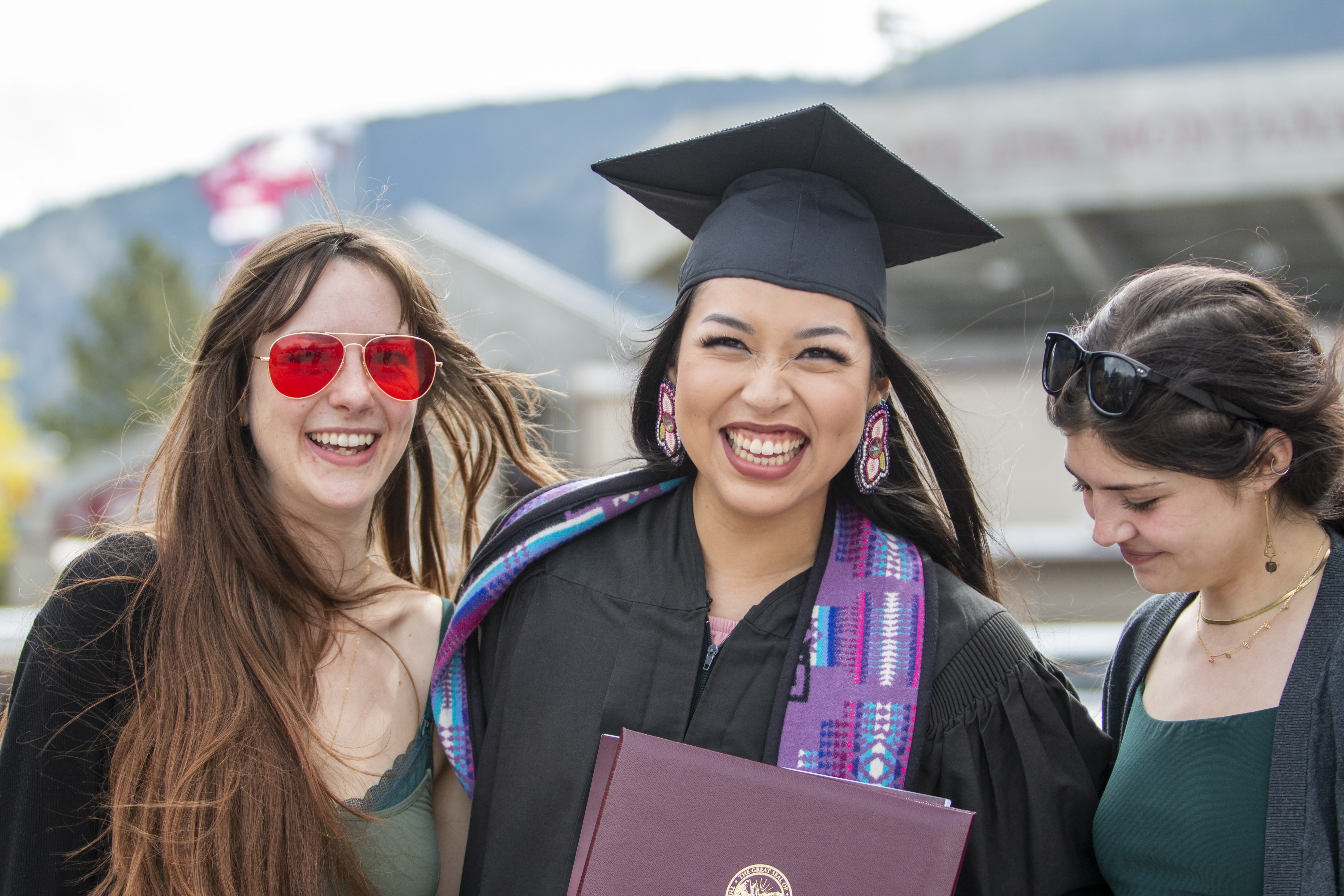 Three students smiling, the person in the middle wears a black graduation cap and gown.