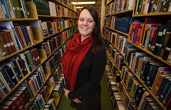 A smiling woman standing confidently between bookshelves filled with books in a library.