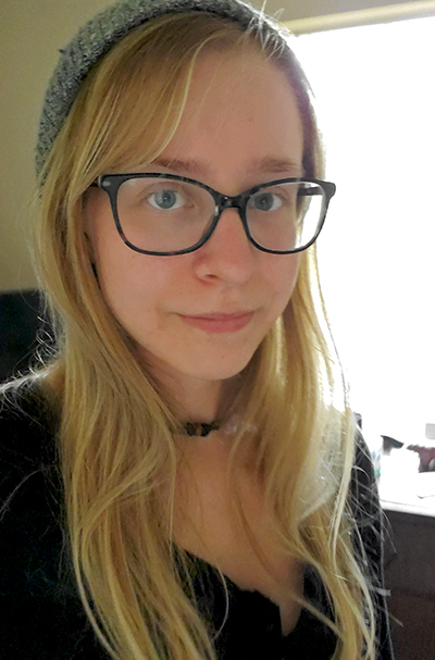 A young woman with long blonde hair and glasses wearing a gray beanie, posing indoors with soft natural light in the background.