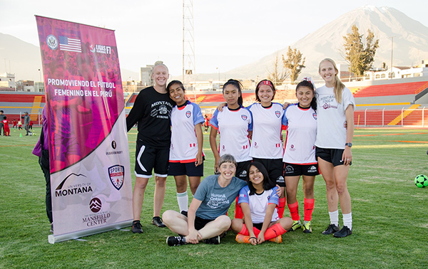 A soccer team photo of five young Peruvian women and three older American soccer players standing and sitting in a soccer field 