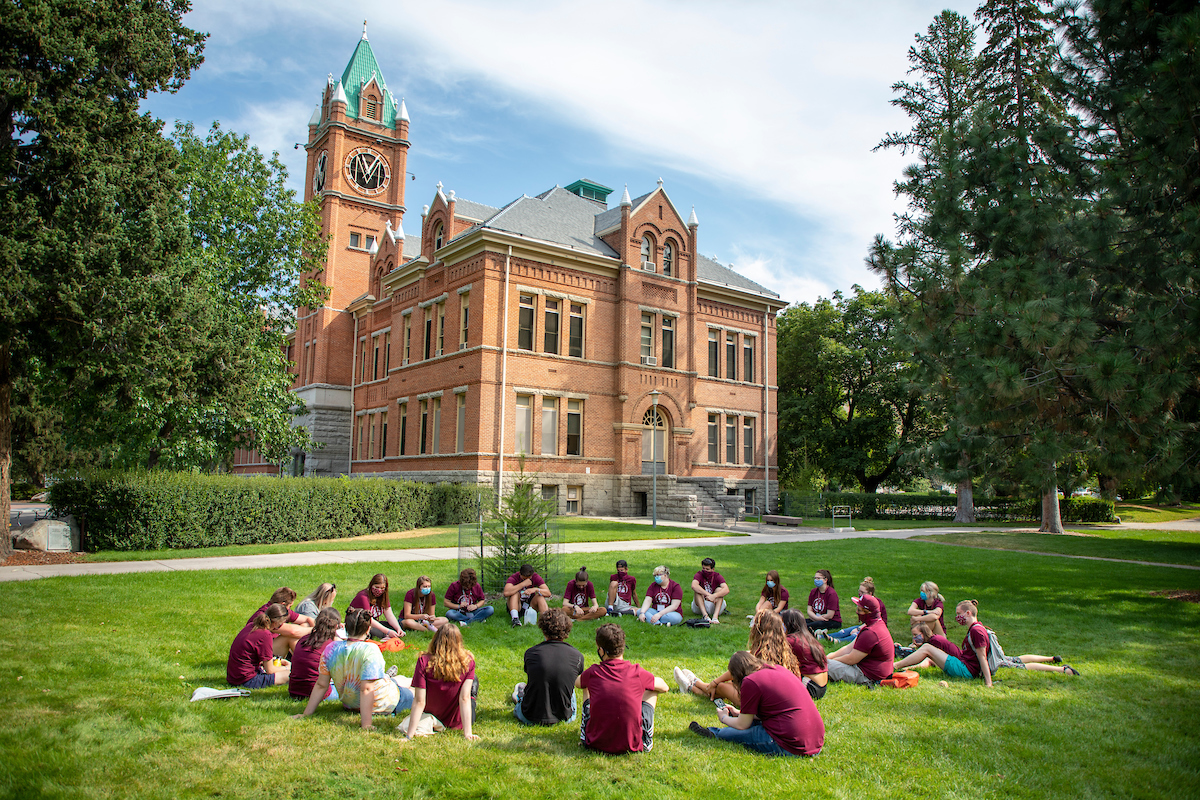 A group of students wearing maroon sits in a circle on the grass with Main Hall in the background 