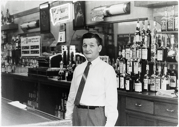 An old photo of a man in a white collar shirt standing in front of a bar 