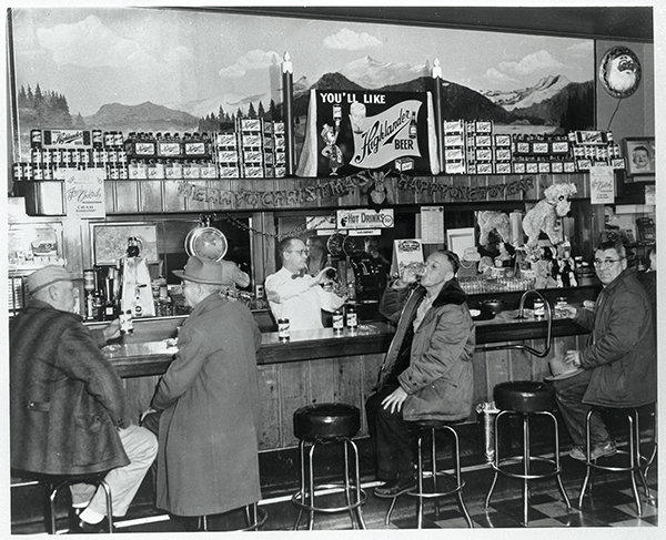 An old image of four men at a bar with a bartender in the background, stacks of bottles and a sign that says You'll Like Highlander Beer 