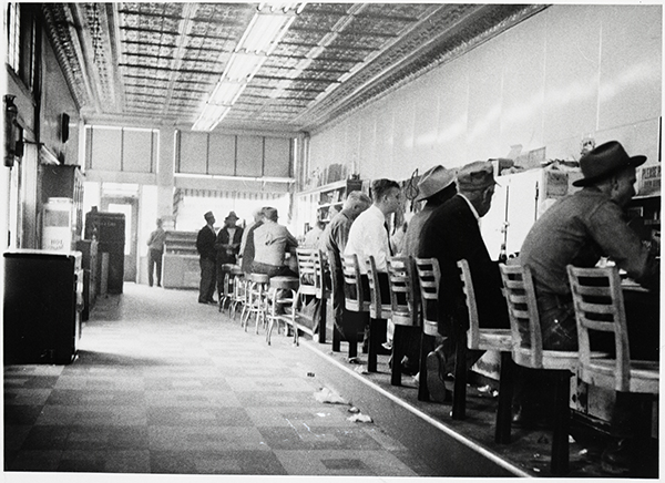 An old image of men lined up at a bar on barstools against the wall 