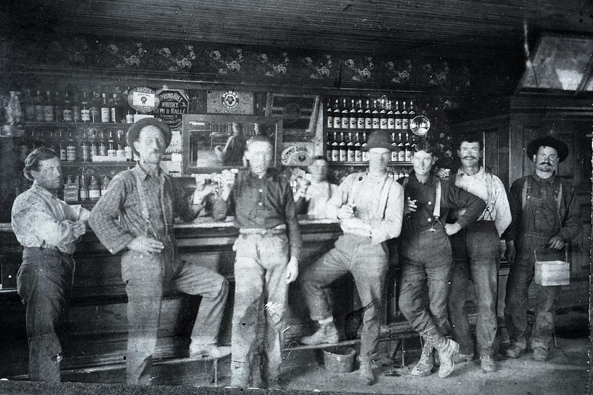 This is a very old photo of seven men standing in front of a saloon bar with bottles in the background 