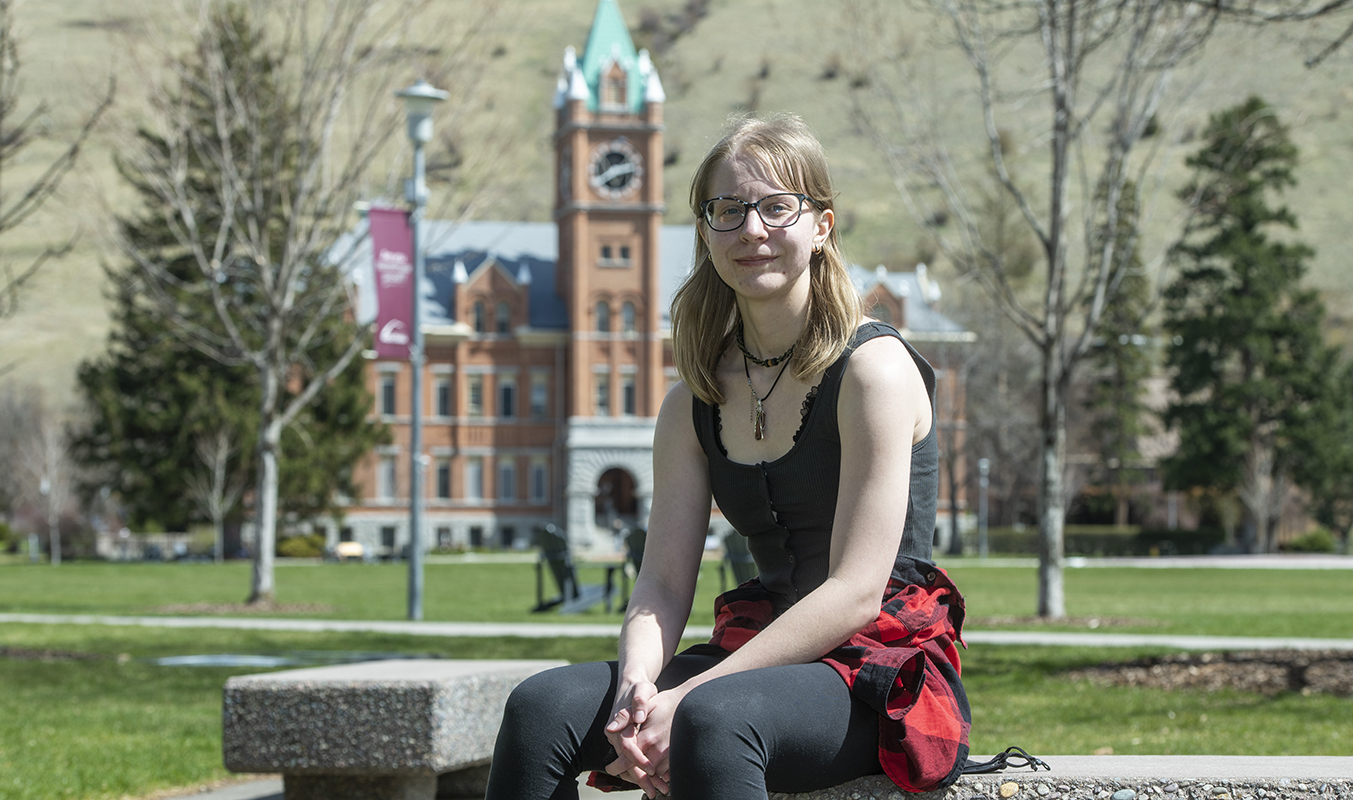 An image of a blonde-haired girl with glasses sitting on a bench with Main Hall blurred out in the background 