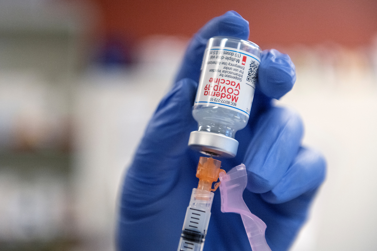 A gloved hand holds a bottle that says Moderna COVID-19 vaccine with a syringe coming out the bottom 