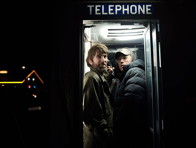 Actor Sharlto Copley and director Tony Stone pose in cramped phone booth at night.