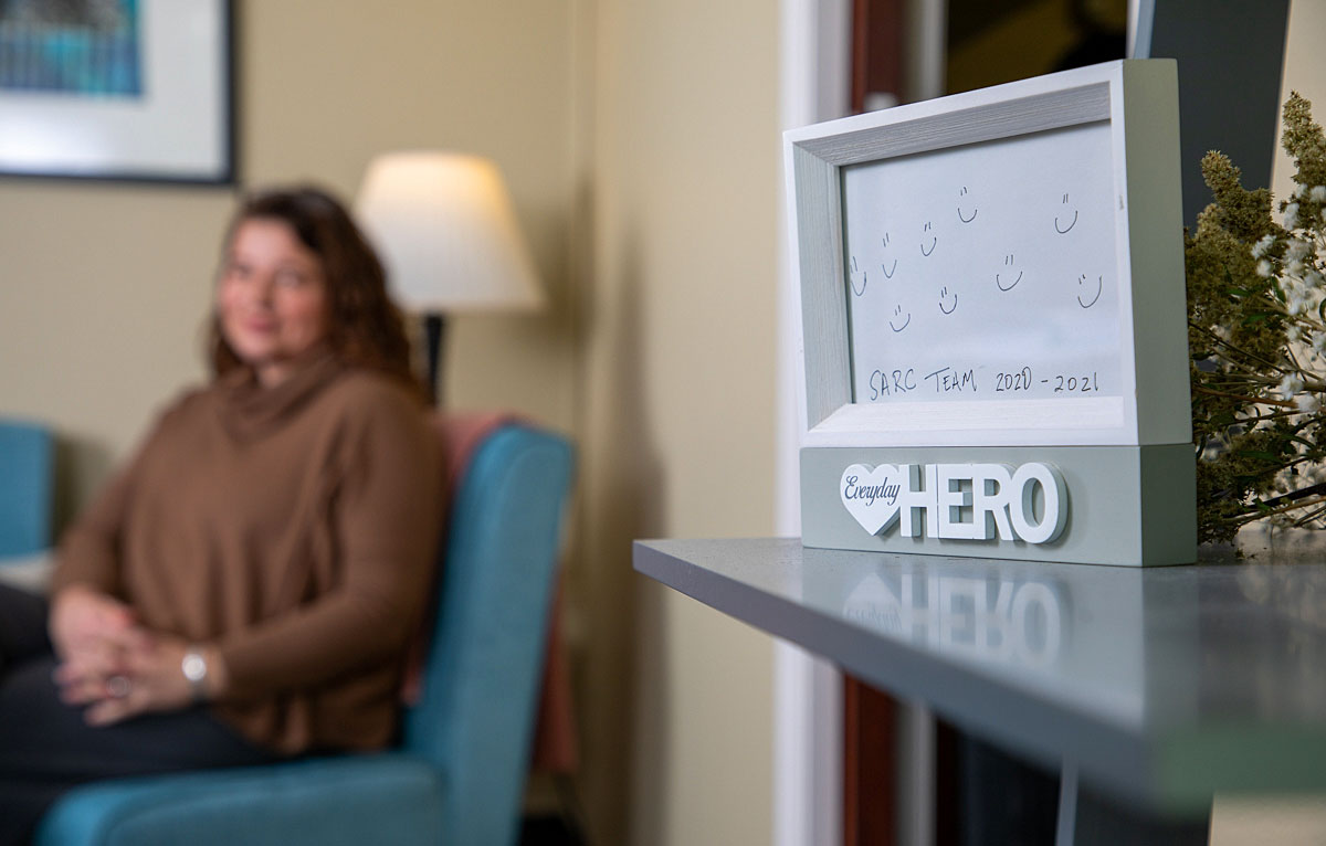 A picture of Jen Euell in her office near a picture that says "Everyday Hero:SARC Team 2020-21"