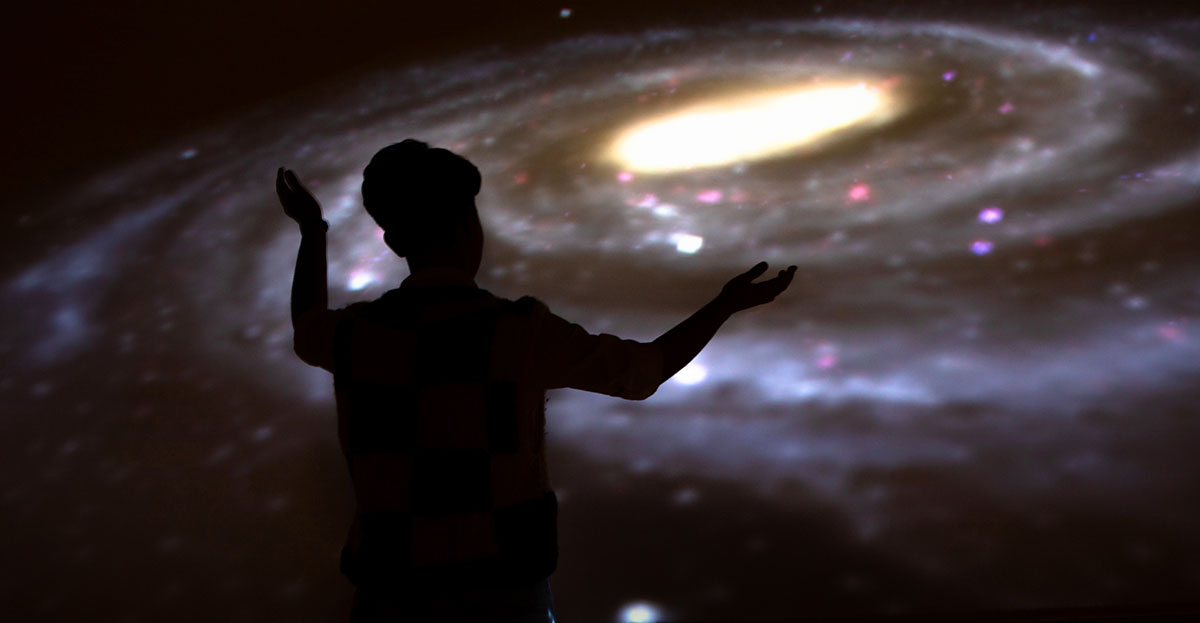 silhouette of a person gesturing in front of a large image of a spiral galaxy