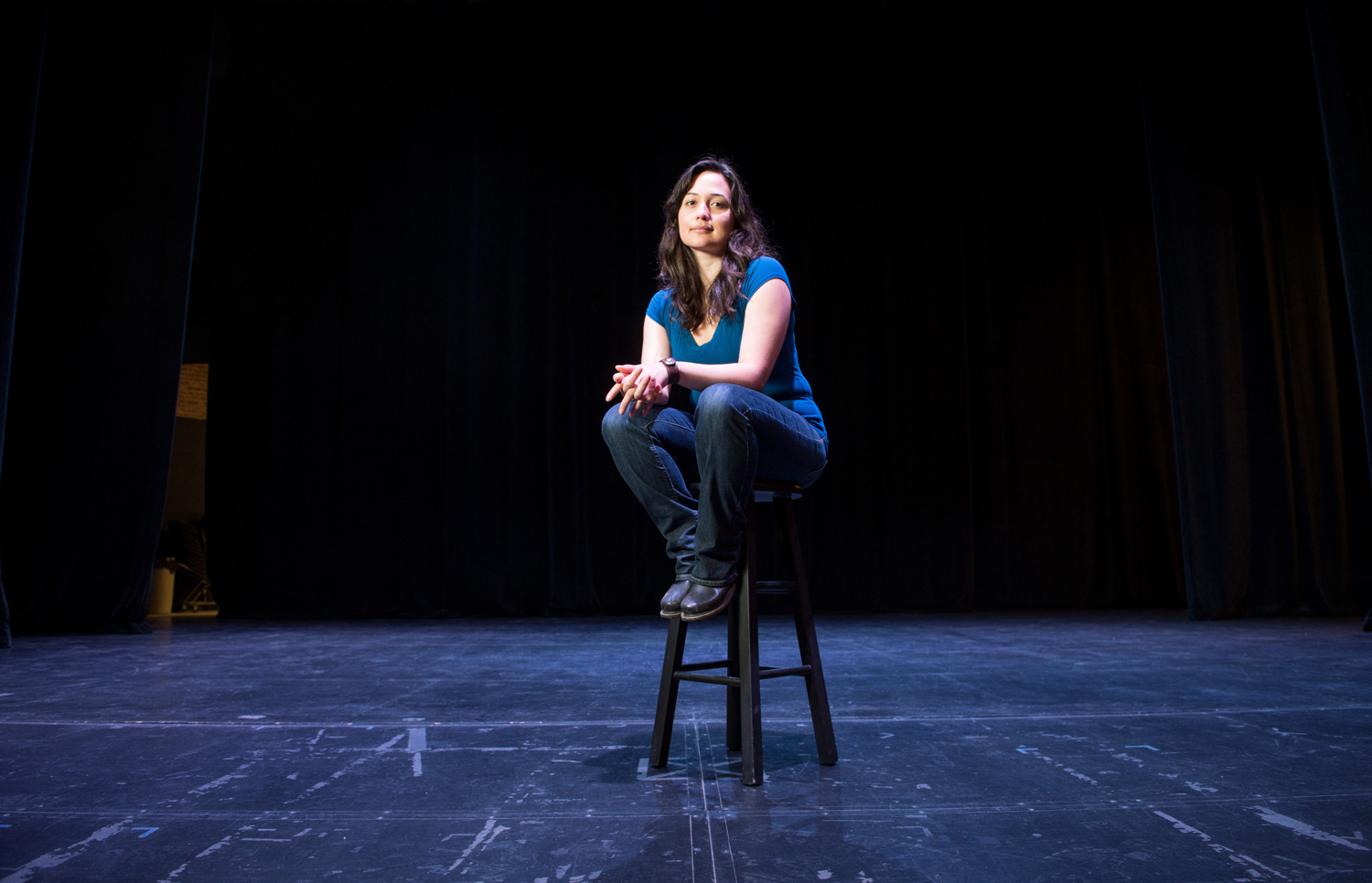 An image of Lily Gladstone sitting on a stool on stage. 