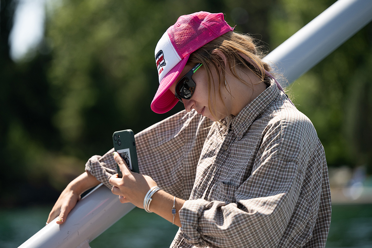 A person wearing a pink cap and sunglasses is looking at their phone while leaning against a railing outdoors.