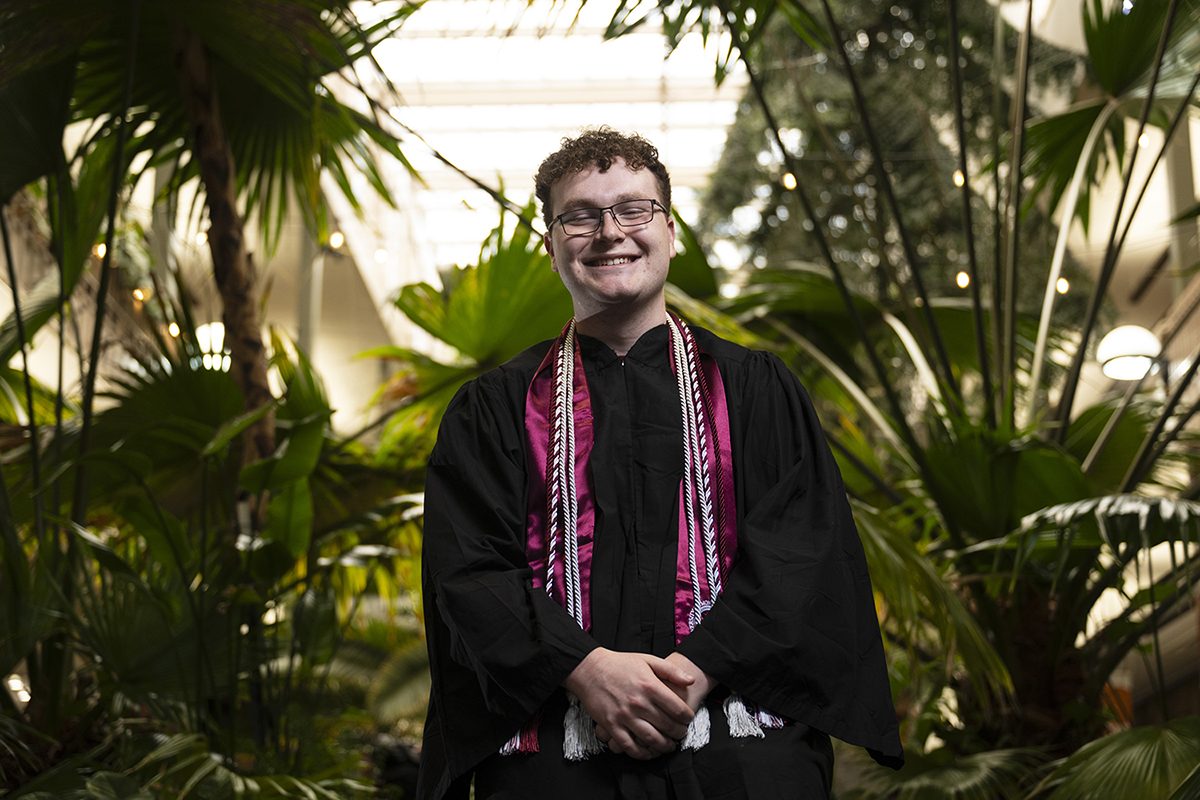 A picture of Liam Edwards in his graduation regalia by the jungle plants of the University Center.