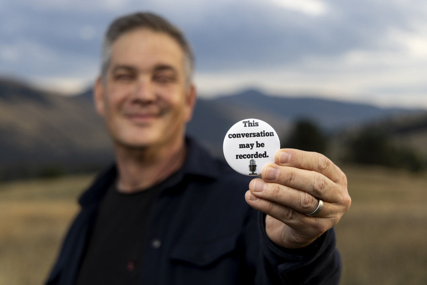 An image of Stephen Yoshimura holding a button that says "this conversation may be recorded."