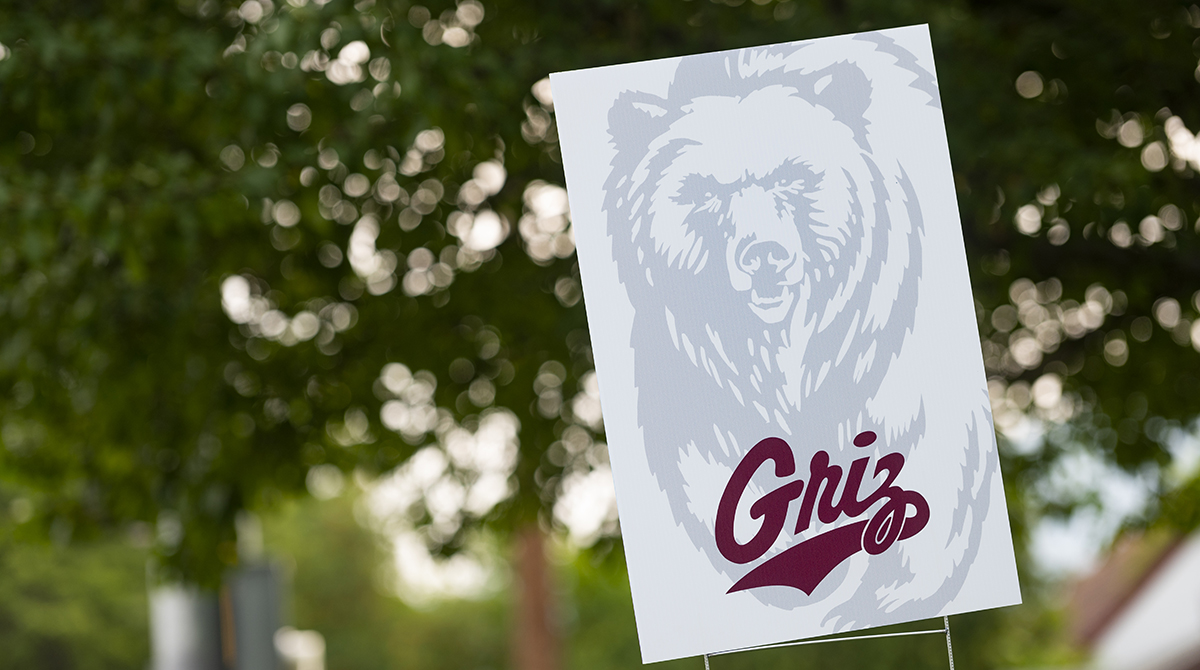 A sign with a Grizzly bear sits in front of tree in town 