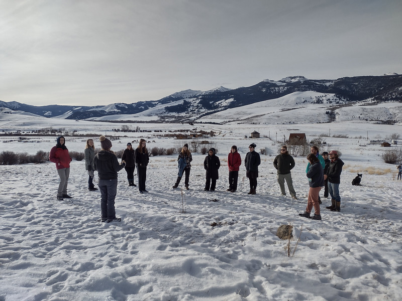A group of people gathered in an open snowy landscape, with mountains in the background, participating in an outdoor discussion or workshop.