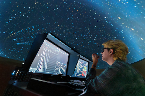 student sits in front of two computer screens with the planetarium dome displaying a starry sky in the background.