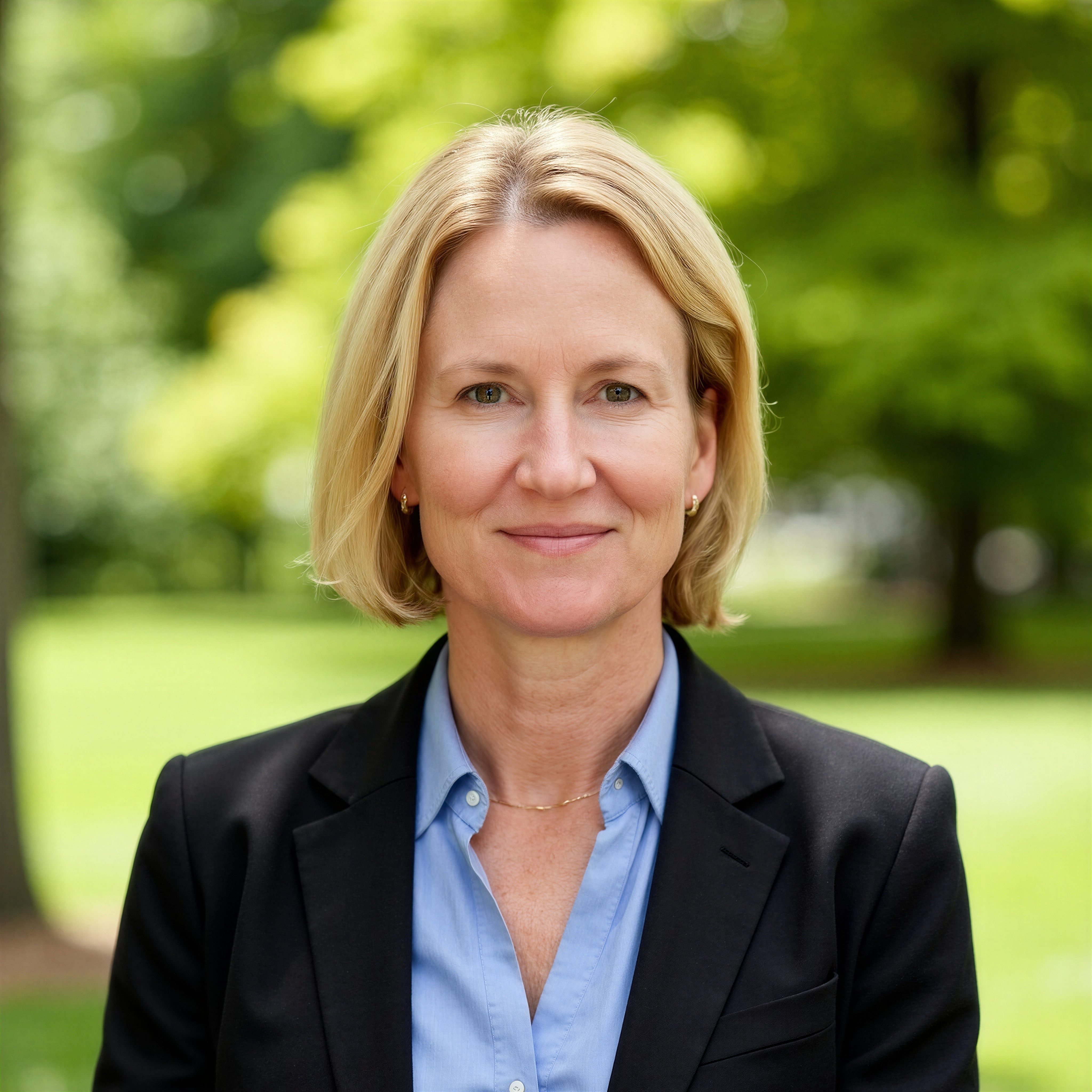 Head-and-shoulders portrait of a blonde woman in a blazer and blue shirt, standing outdoors with a blurred green background.
