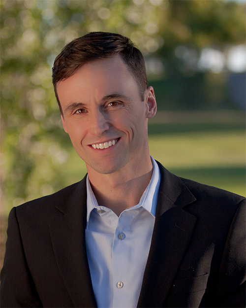 Smiling Brandon Rennie in a suit jacket and light blue shirt outdoors with a blurred green background.